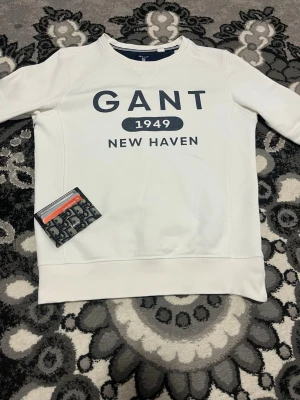 Vit sweatshirt från GANT - Vit sweatshirt från GANT med trycket 'GANT 1949 NEW HAVEN' i mörkblått på bröstet. Tröjan har rund halsringning och ribbade muddar vid ärmslut och nederkant. Perfekt för en clean och klassisk stil.