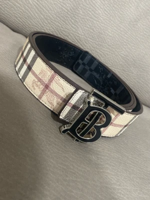 Burberry bälte med rutigt mönster - Snyggt Burberry-bälte med klassiskt beige, svart och rött rutmönster. Bältet har en stor metallspänne med Burberrys ikoniska B-logga framtill. Tillverkat i läder med tygdetaljer och passar perfekt för att lyfta vilken outfit som helst. Helt ny 