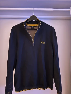 Svart brun/beige tröja från Hugo Boss - Snygg svart långärmad tröja från Hugo Boss med gul logga på bröstet och gul detalj vid krage och nederkant. Tröjan har half zip och ribbade muddar. Perfekt för dig som gillar stilrena och skön stil.