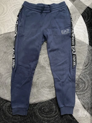 EA7 Emporio Armani träningsbyxor joggers mörkblå storlek S - Säljer mina snygga EA7 Emporio Armani träningsbyxor / joggers i mörkblå färg (navy). Modellen har klassiska stora vita loggor längs båda sidorna på benen + EA7-logga på låret. Supertrendiga just nu! •  Storlek S  •  Fint skick Helt äkta från Emporio Armani EA7-serien.