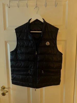 Svart dunväst från Moncler - Snygg svart dunväst från Moncler med klassisk quiltad design och Moncler-logga på bröstet. Västen har dragkedja framtill och två fickor med dragkedja. Perfekt för lager-på-lager och ger en clean look till din outfit.