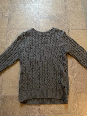 Grå kabelstickad tröja från CRW - Snygg grå kabelstickad tröja från CRW med rund halsringning och ribbade muddar. Tröjan har ett klassiskt flätmönster 