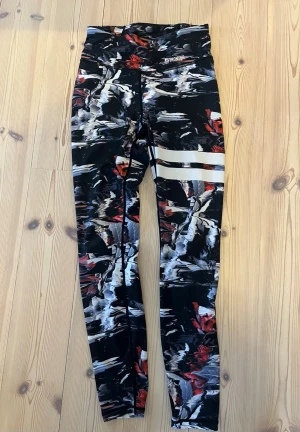 Mönstrade tights från Stronger - Snygga träningstights från Stronger med abstrakt mönster i svart, vitt, rött och grått. Hög midja och två vita ränder på ena benet. Tillverkade i stretchigt funktionsmaterial som passar perfekt för gym eller löpning.