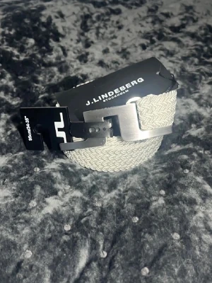 Vitt/beiche flätat bälte från J.Lindeberg - Snyggt beige flätat bälte från J.Lindeberg med silverfärgat spänne och svart läderdetalj vid spännet. Perfekt accessoar för att lyfta din outfit och funkar lika bra till jeans som till chinos. Klassisk design med modern touch.