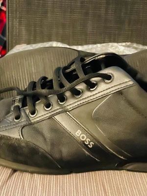 Svarta sneakers från BOSS (42) - Snygga svarta sneakers från BOSS med stilrena detaljer och logga på sidan och sulan. Skorna har snörning, rund tå och är tillverkade i en mix av skinn och textil med inslag av mocka. Perfekta för dig som gillar en clean och modern look. St 42 (Eu) men känns som 42.5/43. Obs. Original Boss Kartong medföljer 