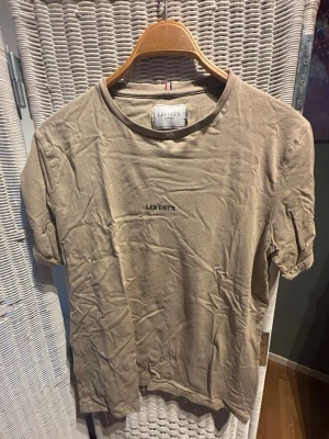 Beige t-shirt från Les Deux storlek M - Säljer en beige t-shirt från Les Deux med diskret logga på bröstet. Klassisk rund halsringning och korta ärmar. Tillverkad i mjuk bomull för en skön känsla. Perfekt till jeans eller shorts för en enkel och clean look.