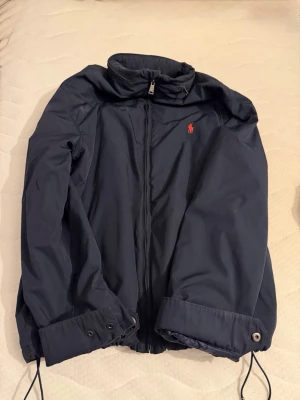 Marinblå vindjacka från Polo Ralph Lauren - Snygg marinblå vindjacka från Polo Ralph Lauren med dragkedja framtill och hög krage. Jackan har det klassiska röda Polo-logotypen broderad på bröstet och justerbara muddar med knappar. Perfekt för blåsiga dagar. Luva går att veckla ut.