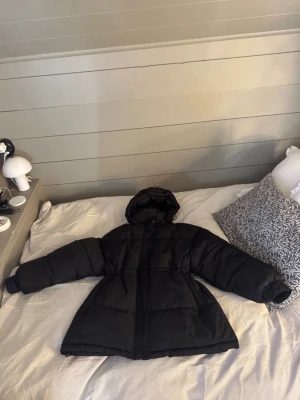 Svart vinterjacka med huva (xs) - Snygg svart pufferjacka från NA-KD med huva och dragkedja framtill. Jackan har en oversized passform, en justerbar midja och långa ärmar, perfekt för kalla dagar. Tillverkad i ett mjukt och vadderat material som håller dig varm hela vintern.                        Använd sparsamt och är i gott skick (något urtvättad i insidan av kragen). Passar Xs/s, är mer som en s i storlek