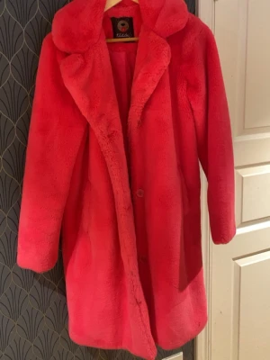 Rosa teddyjacka från Taddy's - Supermjuk och fluffig teddyjacka i stark rosa från Taddy's. Jackan har en klassisk krage, knäppning framtill och långa ärmar. Perfekt för dig som vill sticka ut och hålla dig varm under kyliga dagar.