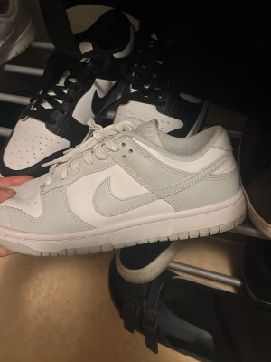 Nike Dunk Low vita sneakers - Nike Dunk Low sneakers i vitt och ljusgrått med klassisk snörning och rund tå. Skorna har en platt sula och är tillverkade i syntet och skinn. Perfekta för dig som gillar stilrena och tidlösa sneakers.