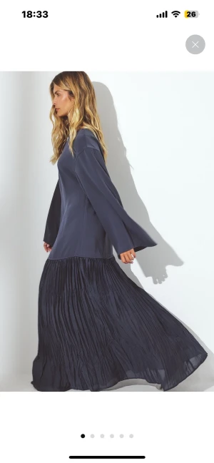 Mörkblå långklänning med plisserad kjol - Säljer en stilren mörkblå långklänning med plisserad kjol och vida långa ärmar. Klänningen har rund halsringning och en enkel knappdetalj i nacken. Den är tillverkad i ett lätt, flowy material som ger ett snyggt fall och extra rörelse till kjolen. Aldrig använd med prislapp. Super fin till bal eller andra festligheter.