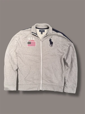 Grå ziptröja från Polo Ralph Lauren - Grå ziptröja från Polo Ralph Lauren med stor mörkblå polospelare broderad på bröstet. Tröjan har USA-flagga och texten 'USA' på vänster sida samt vita detaljer vid dragkedjan och kragen. Tillverkad i mjukt bomullsmaterial och har fickor framtill.