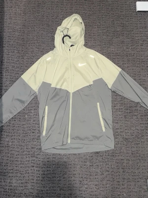 Nike tvåfärgad vindjacka med huva - Snygg vindjacka från Nike i ljusgrått och beige med hel dragkedja och huva. Jackan har två fickor framtill och klassisk Nike-logga på bröstet. Perfekt för blåsiga dagar och streetwear-stil.