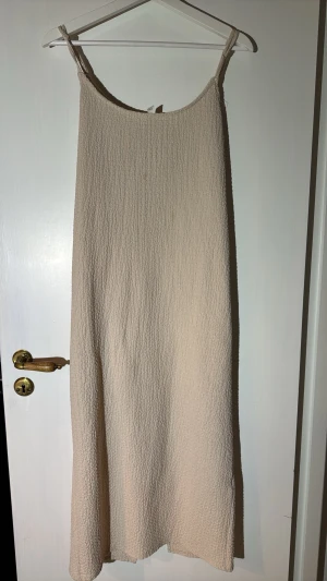 Beige strukturerad klänning L - Minimalistisk beige klänning med smala axelband och strukturerat tyg. Klänningen har en rak passform och är tillverkad i ett lätt, syntetiskt material som ger en skön känsla. Perfekt för dig som gillar enkel och stilren design. Finns två flänckar som syns i vissa ljus.