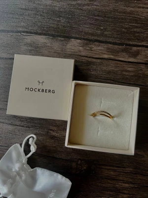 Mockberg Lys Ring – Gold Tone Double Band with Clear Stones - Beautiful Mockberg Lys Ring in a gold-tone finish with an elegant double-band design. One band is decorated with small clear stones, creating a subtle sparkle while keeping a minimalist Scandinavian look.  The ring comes with the original Mockberg box and pouch, as shown in the pictures. Never used and in very good condition.  Original retail price: 599 SEK.  ✨ Perfect for everyday wear, stacking, or as a thoughtful gift.  