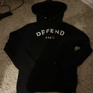 Svart hoodie Defend Paris M - Svart hoodie från Defend Paris i storlek medium. Tröjan har vit text på bröstet, snörning i huvan och klassisk passform. Perfekt för dig som gillar streetwear och vill ha en stilren look med statement-tryck.