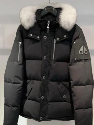 Svart dunjacka med pälshuva från Moose Knuckles - Svart puffer dunjacka från Moose Knuckles med stor vit pälskantad huva, silverfärgade dragkedjor och knappar. Jackan har logotyp på ärmen, flera fickor och quiltade detaljer. Perfekt för kalla vinterdagar och riktigt snygg streetstil.