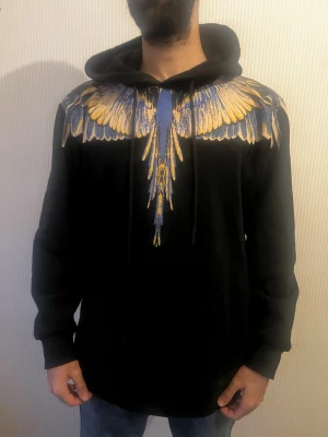 Svart hoodie med färgglada vingar - Säljer en svart hoodie från Marcelo Burlon med ett unikt tryck av blå och gula vingar över axlar och bröst. Hoodien har huva med dragsko och är tillverkad i mjukt bomullsmaterial. Perfekt för dig som vill sticka ut med en cool och kreativ design.