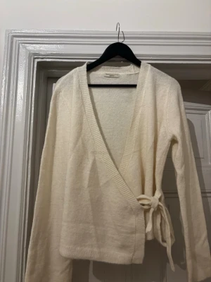 Vit omlottkofta från Gina Tricot - Säljer en mjuk och krämvit stickad omlottkofta från Gina Tricot. Koftan har långa ärmar, ribbade kanter och knyts i sidan med ett band. 