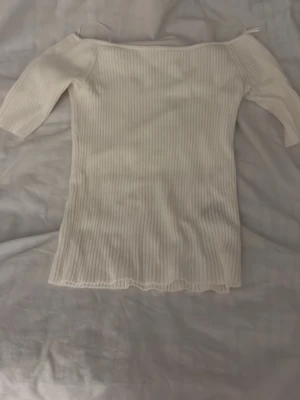 Offshoulder ribbad topp i vitt - Trendig offshoulder topp i vitt med ribbad struktur och korta ärmar. Tunn och stretchig, perfekt för lager på lager eller sommarens outfits. Snygg vågig kant nertill ger en extra detalj. Passar dig som gillar stilrena och enkla plagg.