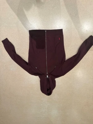 Vinröd hoodie från Superdry - Säljer en vinröd hoodie från Superdry med dragkedja framtill och två stora fickor. Broderad Superdry-logga på bröstet och justerbar huva med snören. Tillverkad i mjuk bomullsblandning, perfekt för chill dagar.