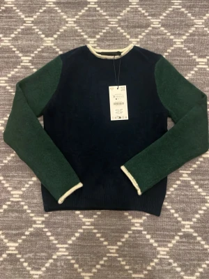 Mörkblå stickad tröja med gröna ärmar - Snygg stickad tröja från Zara i mörkblått med kontrasterande gröna ärmar och ljusa detaljer vid halsringning och ärmslut. Tröjan har en klassisk rund hals och är tillverkad i ett mjukt, värmande material. Perfekt för dig som gillar stilrena färgblock och enkel design.