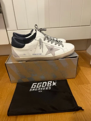 GGDB vita sneakers med grå stjärna - Säljer ett par GGDB sneakers i vitt skinn med grå mockadetaljer och en ikonisk stjärna på sidan. Skorna har grå snörning, mörkblå häl och beige sula. Perfekta för dig som gillar en clean men ändå unik look. Dustbag och originalkartong medföljer. Köpte dem i fel storlek använd fåtal gånger