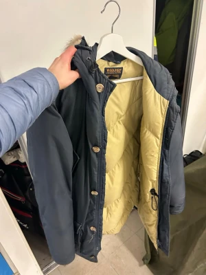 Mörkblå parkas från Woolrich med päls - Säljer en mörkblå parkas från Woolrich med beige quiltat foder och huva med pälsdetalj. Jackan har både dragkedja och knappar framtill samt praktiska fickor. Perfekt för kalla dagar och riktigt snygg till vinterlooken.