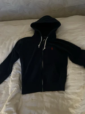 Mörkblå hoodie från Ralph Lauren - Snygg mörkblå hoodie från Ralph Lauren med dragkedja och vit dragsko i huvan. Klassisk röd logga broderad på bröstet. Mjuk bomullskänsla och två fickor framtill. Perfekt för en avslappnad och stilren look. Kan gå ner i pris Passar lite litet på mig som är 169 cm