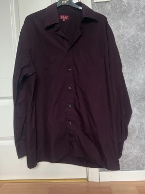 Mörklila skjorta med klassisk krage - Stilren mörklila skjorta med klassisk krage och knäppning framtill. Skjortan har lång ärm och en bröstficka, perfekt för dig som vill ha en enkel men snygg look. Passar till många olika outfits och är lätt att matcha.