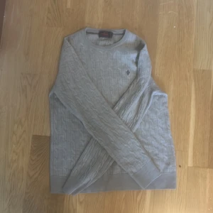 Beige kabelstickad tröja Morris i storlek m men passar s/xs - Snygg beige kabelstickad tröja från Morris i 100% merinoull. Rund hals och diskret Morris-logga broderad på bröstet. Perfekt för dig som gillar klassisk stil med en modern touch. Långärmad och mjuk, passar till jeans eller chinos.