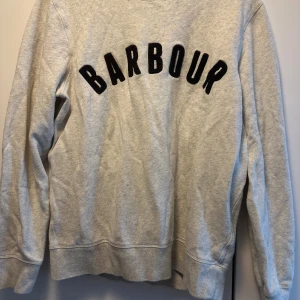 Grå sweatshirt från Barbour - Säljer en grå sweatshirt från Barbour med svart text i ett coolt typsnitt över bröstet. Tröjan har långärmade ärmar och en klassisk rund hals. 