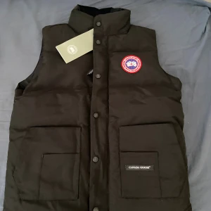 Svart dunväst från Canada Goose - Svart dunväst från Canada Goose med hög krage, två stora fickor framtill och klassisk logotyp-patch på bröstet. Västen har tryckknappar och är tillverkad i slitstarkt material, perfekt för kyliga dagar.
