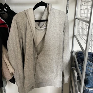Grå hoodie med dragkedja från Weekday - Säljer en ljusgrå hoodie från Weekday med hel dragkedja framtill, huva med snörning och två stora fickor. Tröjan är i mjuk bomullsmix och har långa ärmar. Perfekt för chill dagar och enkel att matcha med jeans eller joggers.