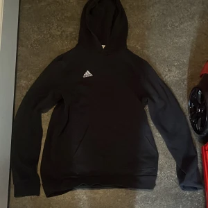 Svart Adidas hoodie med luva - Svart hoodie från Adidas med klassisk logga på bröstet. Tröjan har huva, långa ärmar och ribbade muddar vid ärmslut och nederkant. Perfekt för chill eller träning, enkel och stilren design.