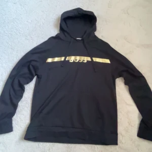 Svart hoodie från BOSS med guldtryck - Svart hoodie från BOSS Homewear i storlek M. Tröjan har huva med snörning och ett stilrent guldtryck med BOSS-logga över bröstet. Långärmad och tillverkad i mjukt material som är skönt att ha på sig. Perfekt för chill dagar eller när du vill ha en snygg streetstyle.