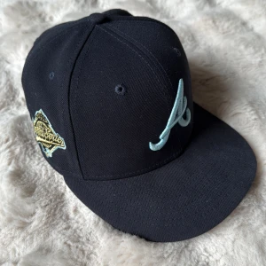 Atlanta Braves 59FIFTY keps New Era - Snygg svart Atlanta Braves 59FIFTY keps från New Era med ljusblå broderad logga framtill och guldfärgad World Series-märke på sidan. Skärmen är svart ovanpå och har en unik ljusblå insida. Klassisk basebollstil i bomull.