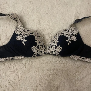Mörkblå spets-bh med broderi - Säljer en mörkblå bh med vacker ljus spets och broderade blommor över kuporna. Bh:n har justerbara axelband och hakknäppning bak. Materialet är mjukt och bekvämt, perfekt för dig som gillar en feminin och elegant look.