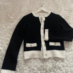 Svartvit kofta med knappar - Stilren svart kofta med vita ribbade detaljer vid krage, ärmslut och nederkant. Koftan har dekorativa fickor framtill och stängs med snygga knappar. Perfekt för dig som gillar klassisk och trendig stil.