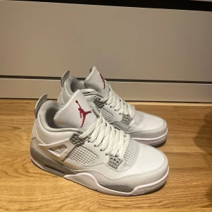 Nike Air Jordan 4 vit/grå sneakers - Nike Air Jordan 4 sneakers i vitt och grått med röda Jumpman-loggor på plösen. Skorna har snörning, meshpaneler och detaljer i skinn samt en platt sula med grå stänk. Klassisk siluett med högt skaft och rund tå, perfekt för dig som gillar streetwear.