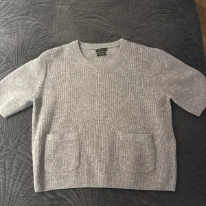 Grå ribbad stickad tröja med fickor - Säljer en grå stickad tröja från Massimo Dutti med ribbad struktur och två praktiska fickor framtill. Tröjan har korta ärmar och rund hals, perfekt för lager-på-lager under kyliga dagar. Enkel och stilren design som passar till allt. Ull blandat med kashmir❤️❤️❤️