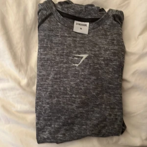 Gråmelerad Gymshark träningströja M - Säljer en gråmelerad kortärmad träningströja från Gymshark i storlek M. Tröjan har svart-vita inslag för att markera dina muskler framförallt axlar och rygg, sportig passform och Gymshark-logga på bröstet som är i rubber material. Perfekt för gymmet eller löprundan, tillverkad i mjukt och stretchigt syntetmaterial. Aldrig använd då den var förstör 