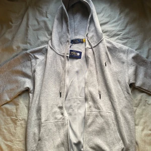Grå hoodie från Polo Ralph Lauren - Snygg grå hoodie från Polo Ralph Lauren med klassisk logga broderad på bröstet. Tröjan har huva med snörning, två stora fickor framtill och en relaxed passform. Perfekt för chill dagar eller när du vill ha en clean och stilren look.