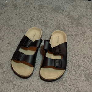 Svarta tofflor från Pier One - Svarta sandaler från Pier One storlek 41, i nyskick med två breda remmar i syntet och justerbara spännen. Fotbädden är beige och har en skön, anatomisk form. Perfekta för sommaren och riktigt chill stil. 