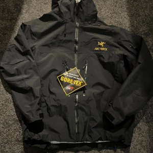Svart Arc'teryx Gore-Tex skaljacka XL - Arc'teryx skaljacka i svart med GORE-TEX-material, perfekt för regniga och blåsiga dagar. Jackan har huva, hel dragkedja, flera fickor och justerbara ärmslut. Snygg gul Arc'teryx-logga på bröstet och minimalistisk design. Helt ny aldrig använd med tags kvar.