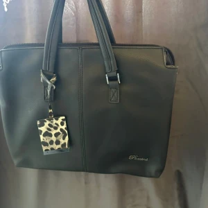 Svart datorväska med leoparddetalj - Stilren svart datorväska från Picard i skinnimitation med dubbla handtag och dragkedja. Väskan har ett snyggt leopardmönstrat fack på sidan och blommigt foder inuti. Perfekt för laptop och har flera praktiska innerfack.