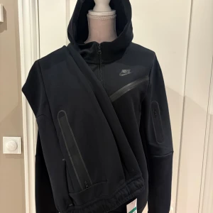 Svart Nike hoodie med dragkedja XL - Svart hoodie och byxor från Nike tech i storlek XL med hel dragkedja och huva. Snygga detaljer med svarta zippar på fickorna och diskret Nike-logga på bröstet. Perfekt för en sportig och chill stil. Materialet är mjukt och känns bekvämt mot huden.