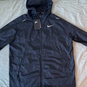 Nike Miler Jacka - Svart vindjacka från Nike med huva och hel dragkedja framtill. Jackan har två sidofickor, meshfoder och reflekterande detaljer på axlarna. Nike-logga på bröstet och Dri-FIT-teknologi för extra komfort. Perfekt för löpning och aktiva dagar.