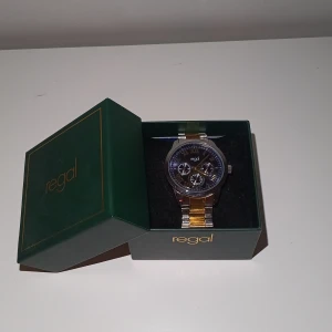 Regal herrklocka i silver och guld - Snygg herrklocka från Regal med blå urtavla och romerska siffror. Armbandet är i metall med både silver- och guldfärgade länkar. Klockan har tre mindre urtavlor och levereras i original grön ask med extra länkar. Perfekt accessoar för dig som gillar stilrena detaljer.
