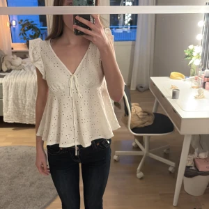 Vit blus med spets och volang - Säljer en vit blus med broderade hålmönster, volangärmar och v-ringning. Blusen har knyt framtill och är lätt utsvängd nedtill. 